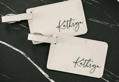 Two beige luggage tags with 'Kathryn' engraved on a dark marble surface