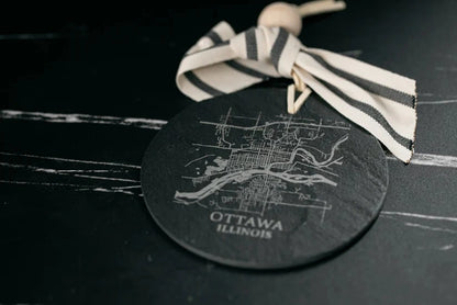Round black coaster with white map design and 'Ottawa, Illinois' text, tied with a striped ribbon on a dark surface.