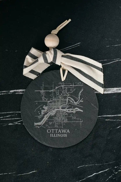 Round black coaster with white map design and 'Ottawa, Illinois' text, tied with a striped ribbon on a dark surface.