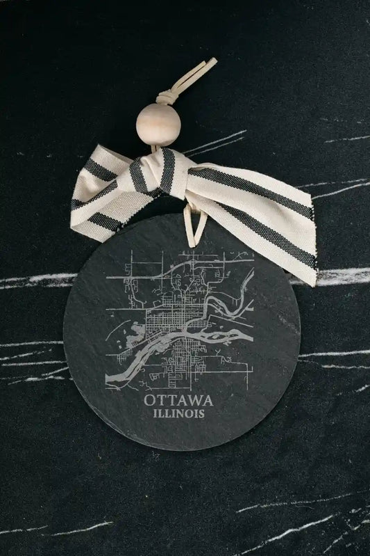 Round black coaster with white map design and 'Ottawa, Illinois' text, tied with a striped ribbon on a dark surface.