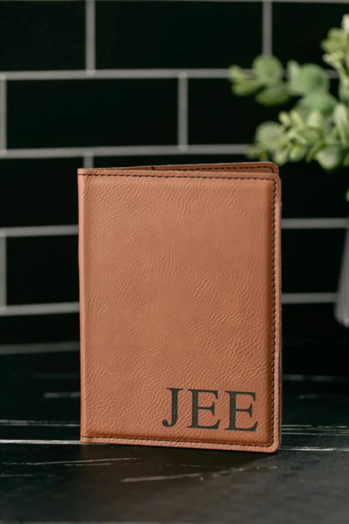 Brown leather wallet with 'JEE' engraving on a dark surface with a blurred green plant in the background.